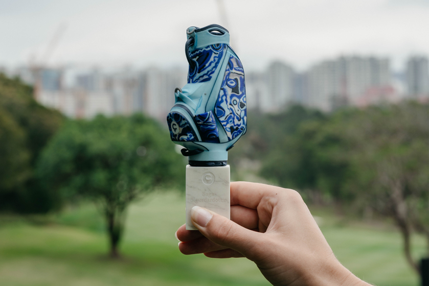 Hand holding a finished miniature golf bag with a blue and teal marbled pattern on a display base, with a city skyline and green trees in the background.