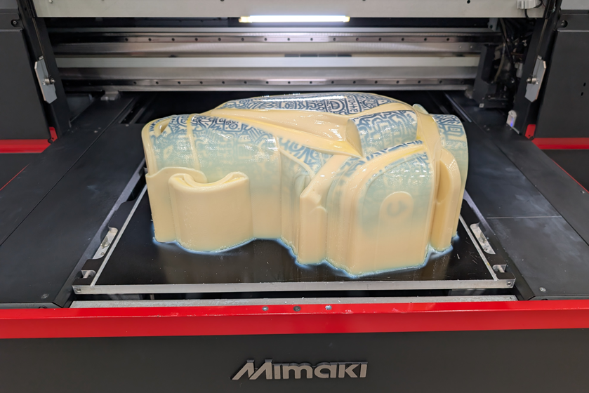 3D printed golf bag model with support material still attached, resting on the build plate of a Mimaki 3D printer, showing a blue and white patterned design partially visible through the wax-like support structure.