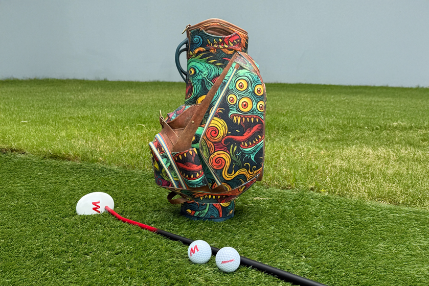 Full-scale Mimaki 3D printed golf bag replica with monster and eye motif design, displayed on a golf course alongside a golf club and balls.